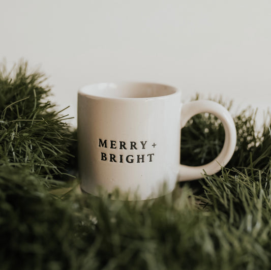 A white stoneware coffee mug with 'Merry + Bright' text on it, placed on a bed of green artificial turf with Christmas decorations.