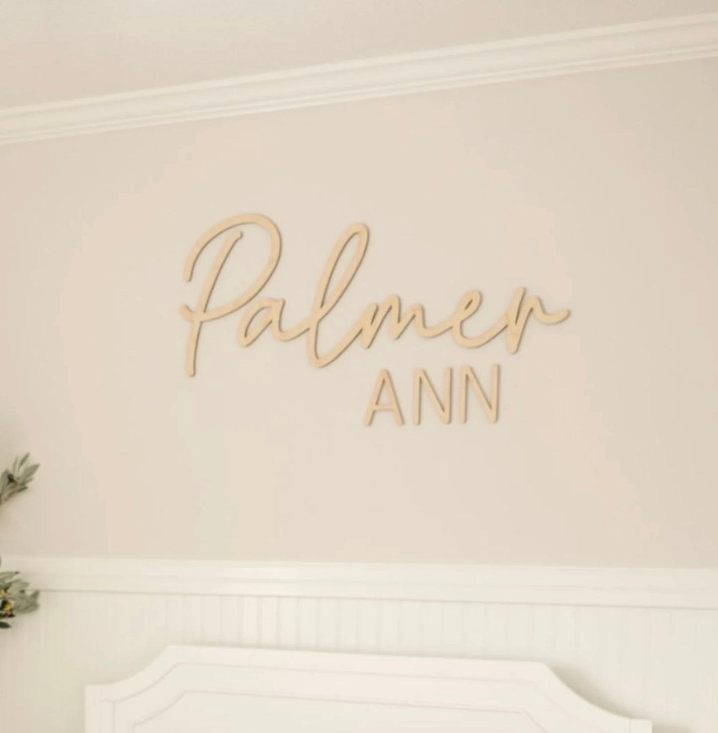 A personalized wooden name sign with 'The Palmer' written in cursive script, placed above a white crib in a nursery room. The room features a pale pink wall, white furniture, and a green plant.