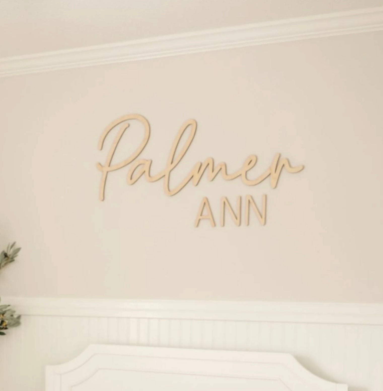 A personalized wooden name sign with 'The Palmer' written in cursive script, placed above a white crib in a nursery room. The room features a pale pink wall, white furniture, and a green plant.