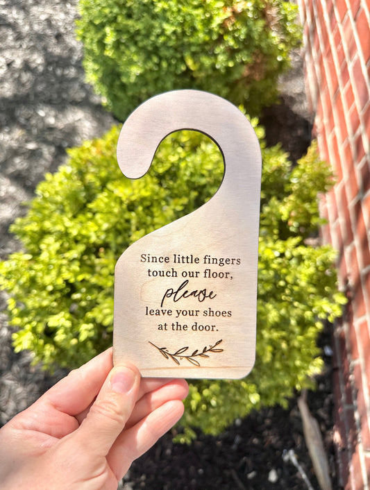 A wooden door hanger with a curved shape and the text 'Since little fingers touch our floor, PLEASE LEAVE YOUR SHOES AT THE DOOR' engraved on it. The hanger is held against a backdrop of greenery.