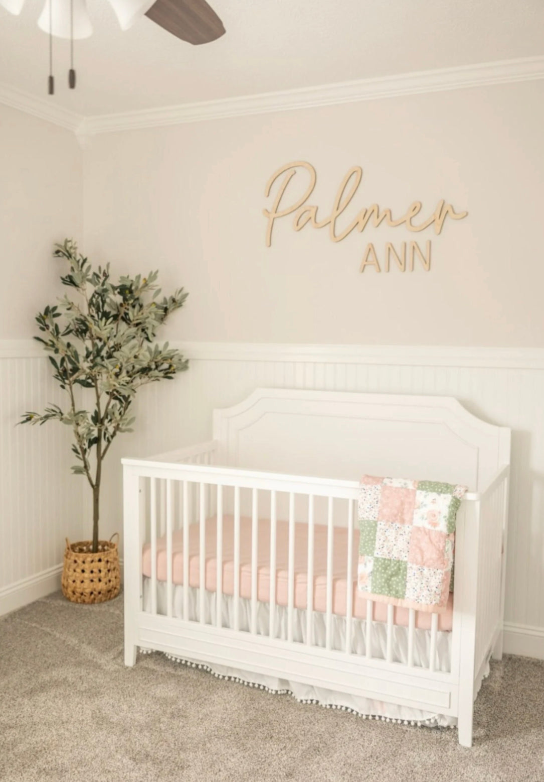 A personalized wooden name sign with 'Palmer Ann' written in cursive script, placed above a white crib in a nursery room. The room features a pale pink wall, white furniture, and a green plant.