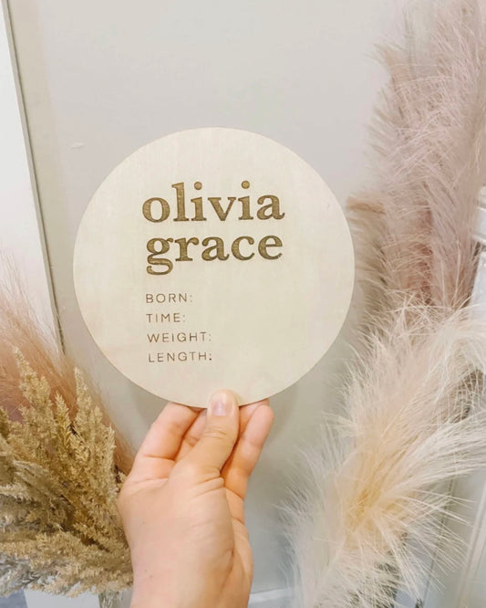 A hand holding a round birch wood plaque with the engraved text 'Olivia Grace' along with the phrases 'Born Time Weight Length'.
