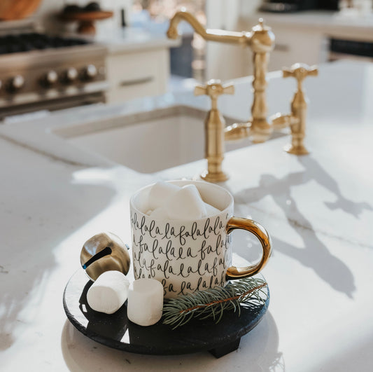 A fine bone china coffee mug with white base and black hand lettering, featuring the phrase 'Fa La La La', displayed on a kitchen counter with gold handle embellishment.