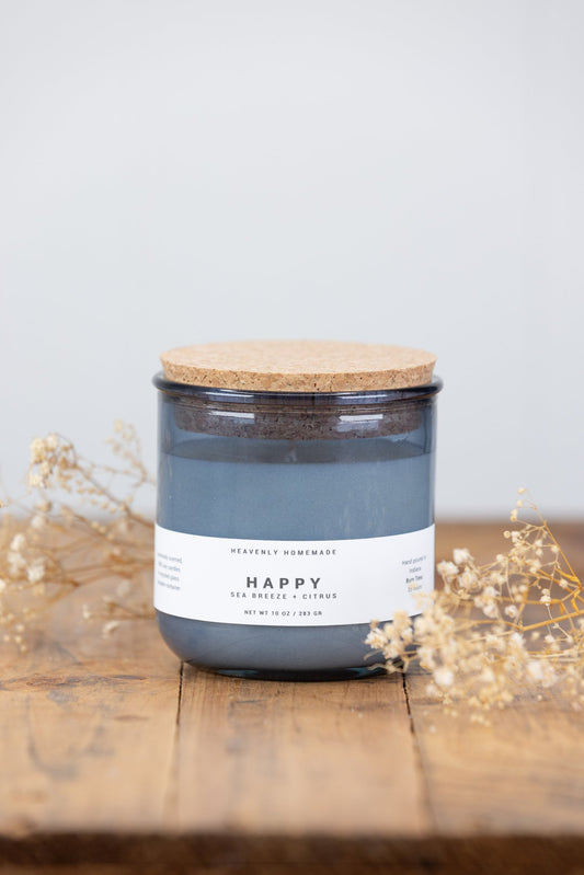 A blue glass jar candle labeled 'Happy' with a wooden lid, placed on a wooden surface with some dried flowers nearby.