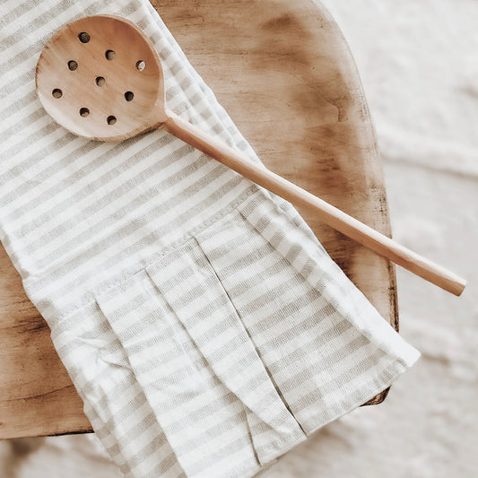A striped hand towel in cream color with tan stripes and a ruffle detail, placed on a wooden surface with a wooden spoon beside it.
