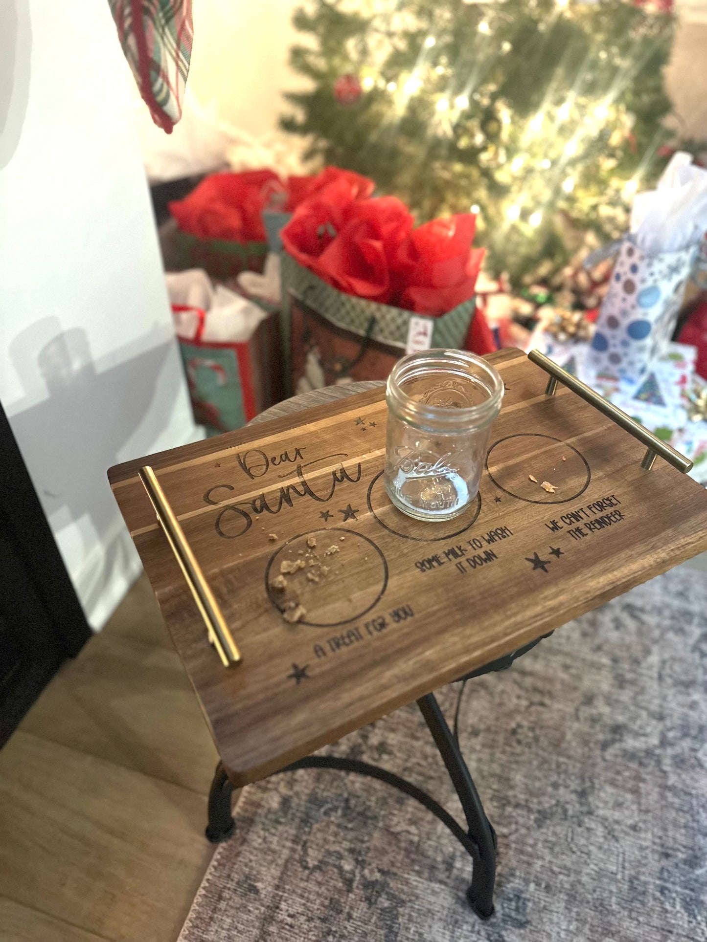 Wooden tray with engraved designs and a glass jar on a surface with a blurred festive background.