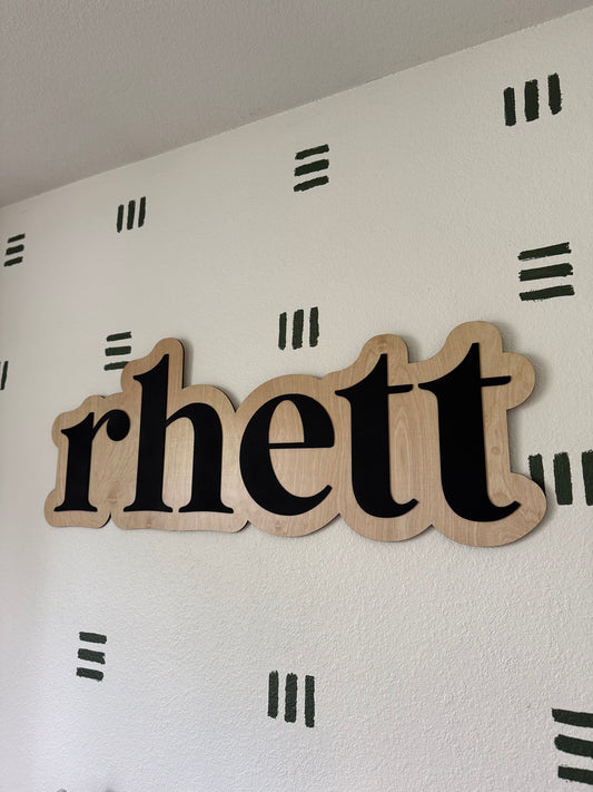 Wooden name sign spelling 'rhett' on a white wall with black patterns