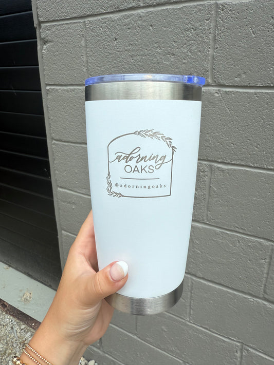 White tumbler with 'Adorning Oaks' branding held against a gray wall.