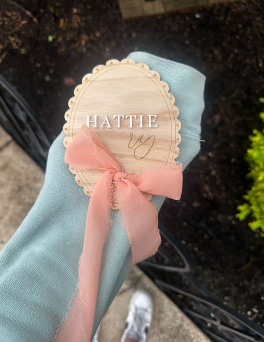 Wooden name tag with 'HATTIE' and a pink ribbon held by a person outdoors.