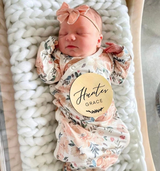 A wooden baby announcement name sign with the engraved text 'Hunter Grace' placed next to a baby girl in a floral-patterned onesie.