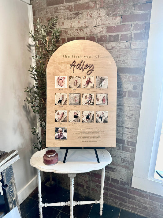 A 3D wooden name sign with laser engraved details, displayed as a birthday party decoration with photos and the name 'Adley' on it, placed on a white table against a brick wall background.