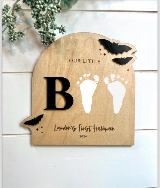 A wooden arch-shaped sign with the word 'BOO' engraved on it, accompanied by a pair of footprints. The sign is designed for capturing a baby's first Halloween, with a black acrylic 3D detail on the pumpkin-shaped cut-out at the top.