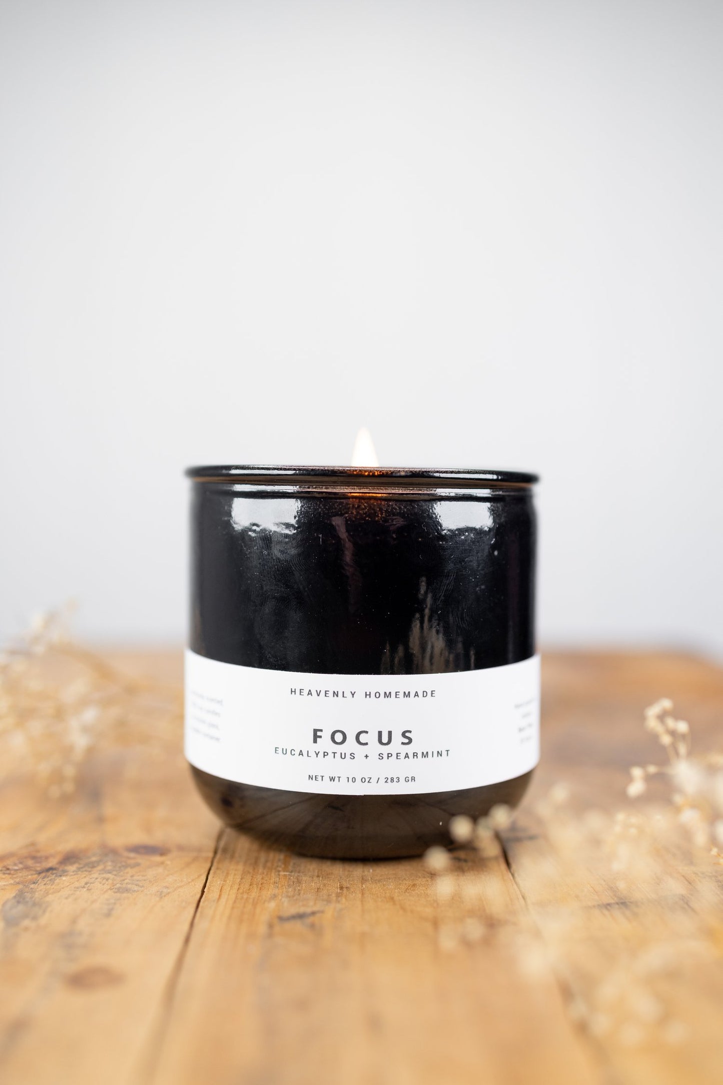 A black jar candle with a cork lid, labeled 'FOCUS' for mood lighting, placed on a wooden surface with dried flowers nearby.