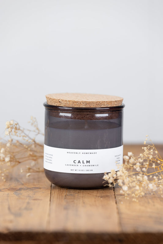 A brown glass jar candle with a label that reads 'Calm - Lavender & Chamomile' on a wooden surface with dried flowers nearby.