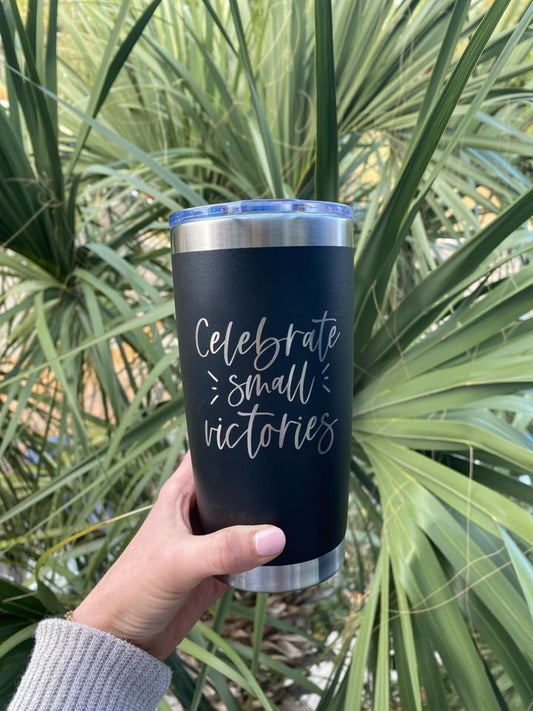 Black tumbler that says Celebrate Small Victories held infront of plant