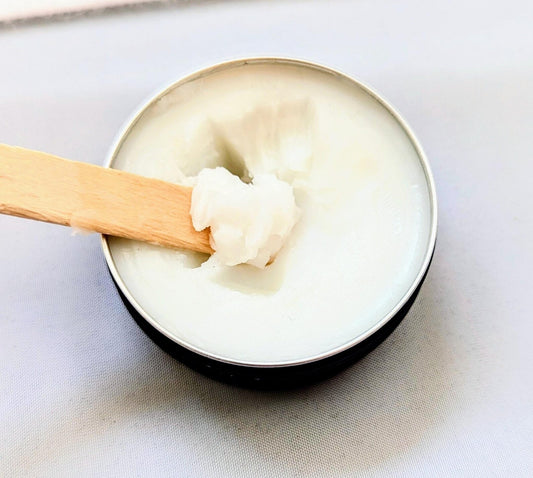 An open container of fragrance balm with a wooden applicator sticking out, showing a solid white cologne paste inside.