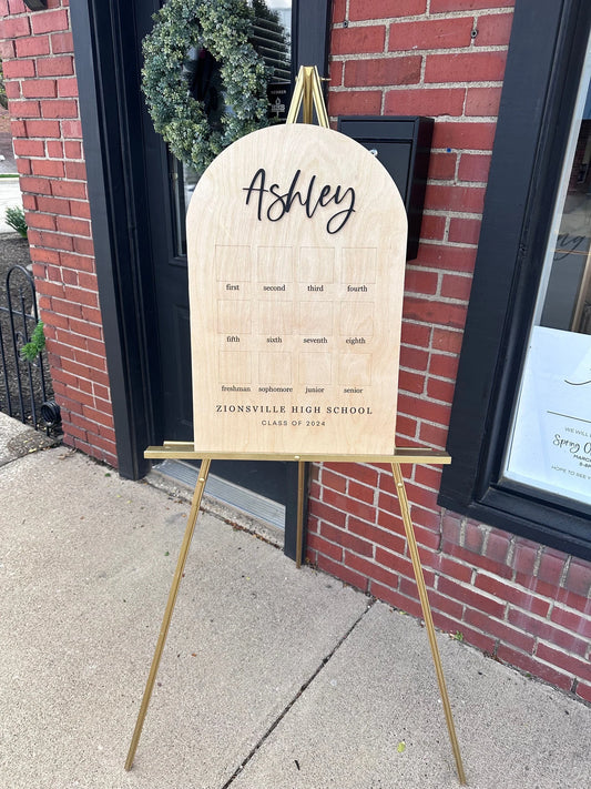 A wooden graduation open house sign with a name written on it, displayed on a stand outdoors.