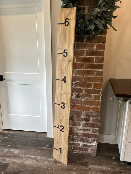 A wooden growth chart with 3D cutout numbers and black painted numbers, standing against a brick wall with a wooden door in the background. The growth chart is taller than the door, which is closed.