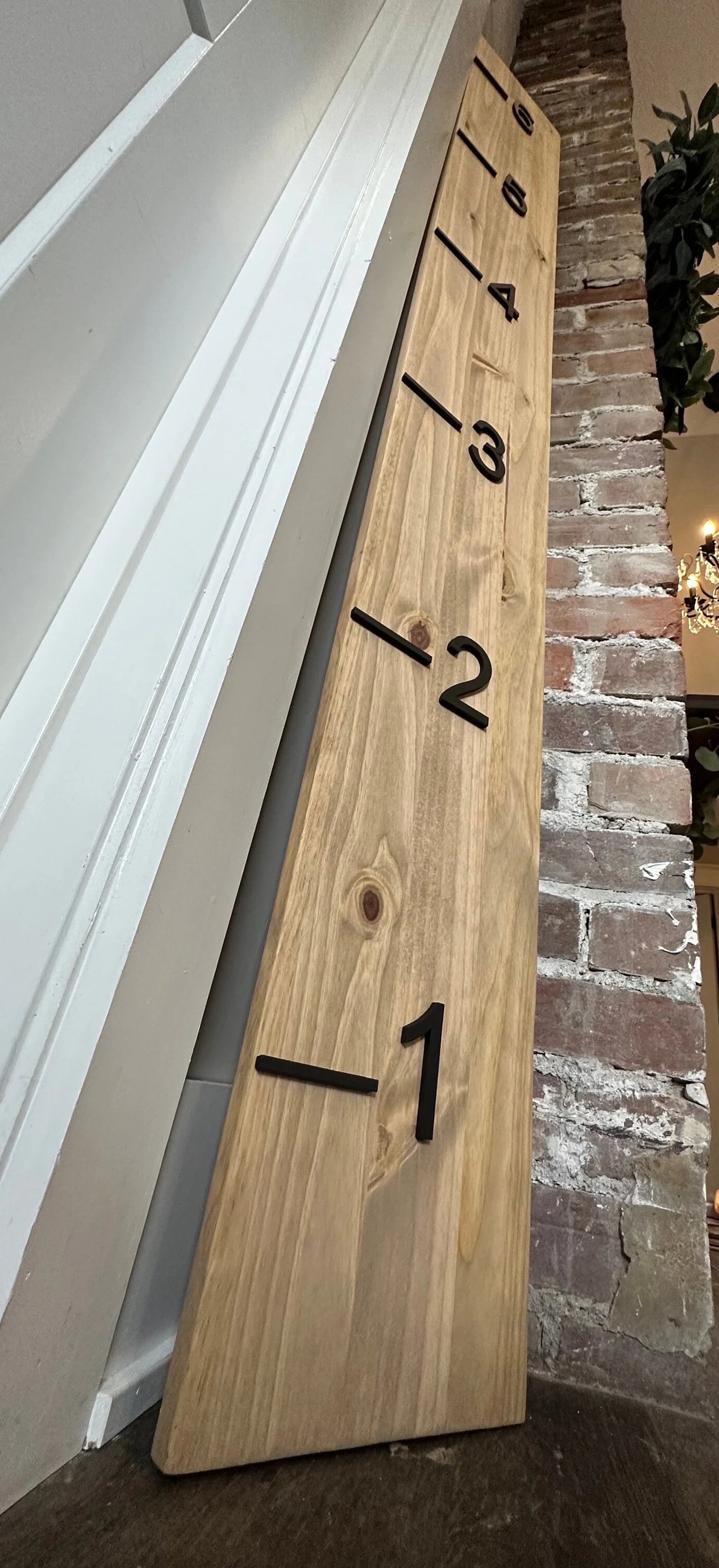 A wooden growth chart with 3D cutout numbers and black painted numbers, standing against a brick wall with a wooden door in the background. The growth chart is taller than the door, which is closed.