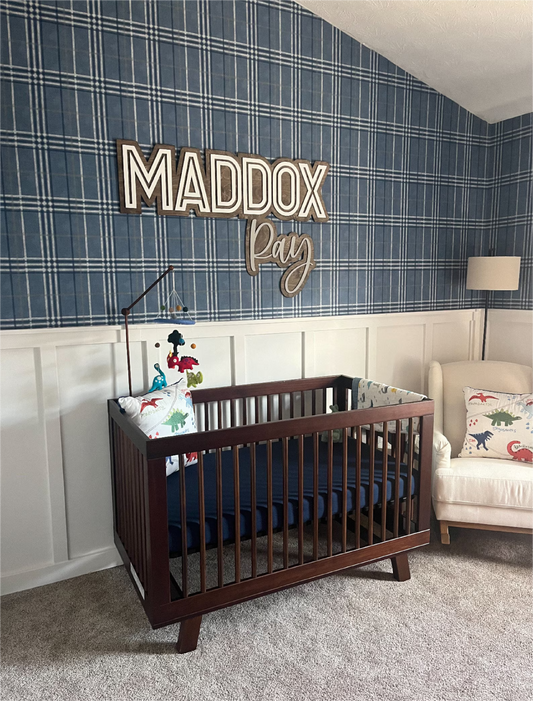 A customized wooden name sign 'MADDOX Ray' is displayed above a wooden crib in a nursery room, with a striped blue and white wall in the background and a white chair to the side.