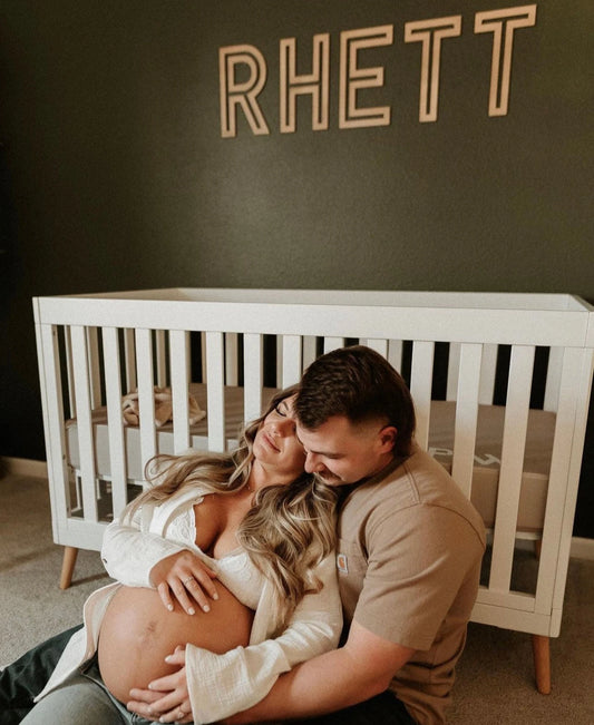 A pregnant woman lying next to a wooden crib with the name 'Rhett' printed on the wall above it.