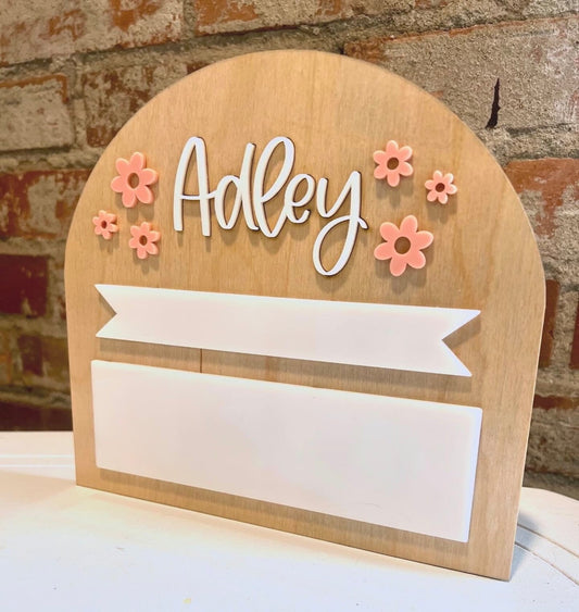 A wooden back to school sign with 'Adley' written on it and decorated with pink and white flowers and a white banner.