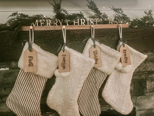Personalized wooden stocking tags with 'Merry Christmas' text and names printed on them, displayed on a wooden rack with Christmas decorations in the background.