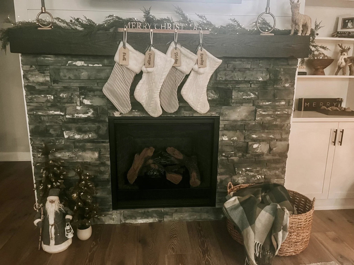 Personalized wooden stocking tags with 'Merry Christmas' text and names printed on them, displayed on a wooden rack with Christmas decorations in the background.