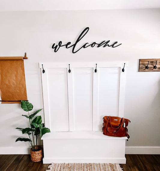 A 'Welcome' wooden cutout in white, mounted on a wall above a brown bench with a brown purse and a green plant, with a black frame on the top.