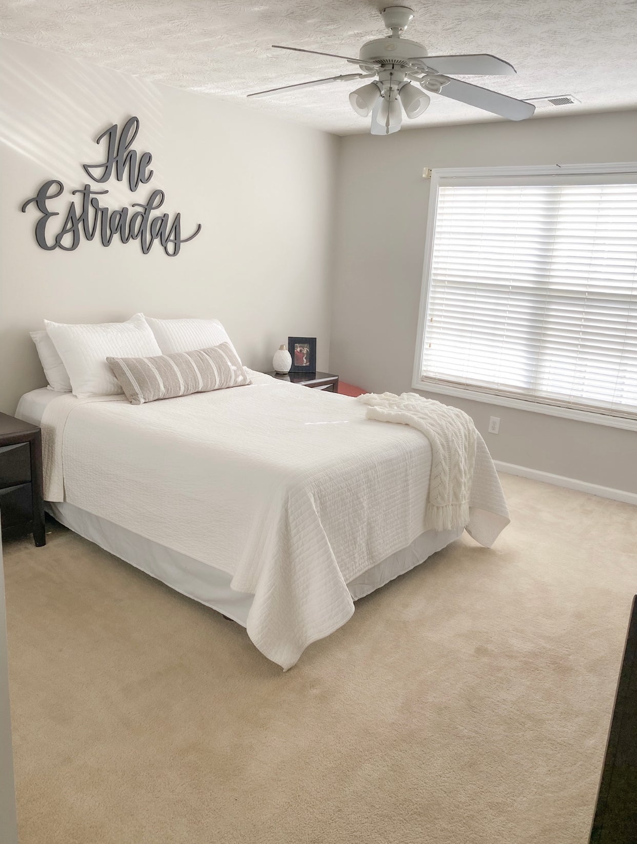 A wooden cutout with the word 'The Estradas' in a large, bold font, mounted on a wall above a bed, with a fan-like ceiling fan in the background.