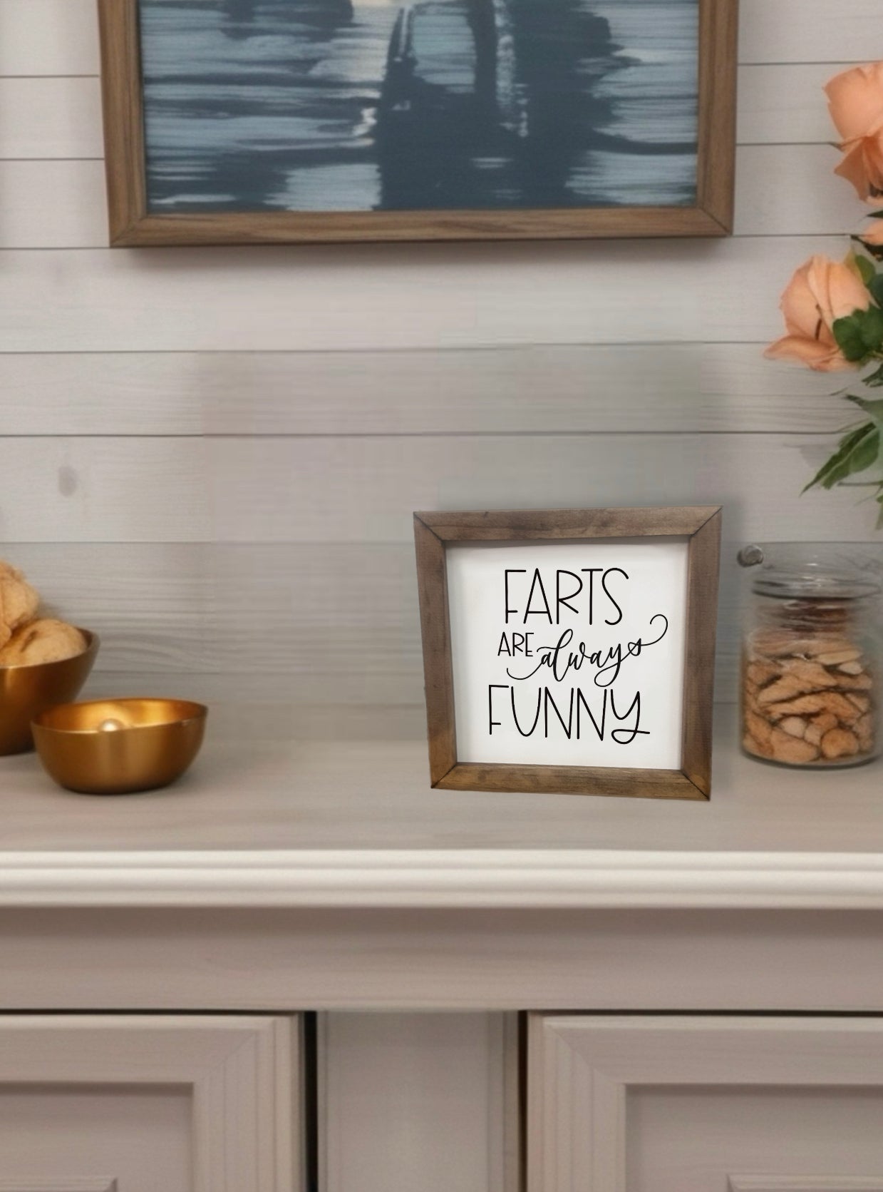 Framed sign with 'Farts are always funny' text on a shelf with decorative items.