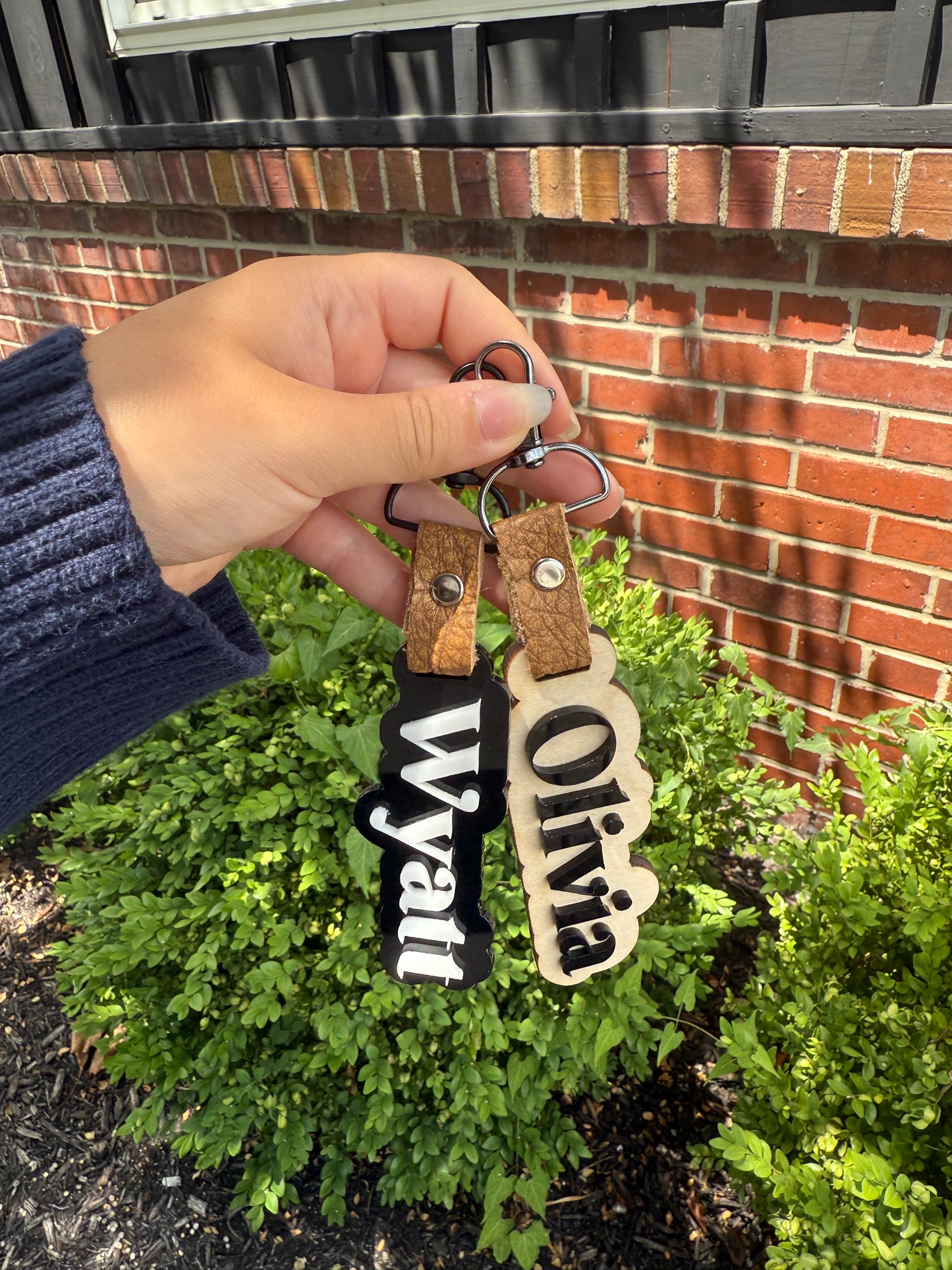Hand holding a wooden name tag with 'Olivia' and 'Wyatt' against a brick wall and green bush background.