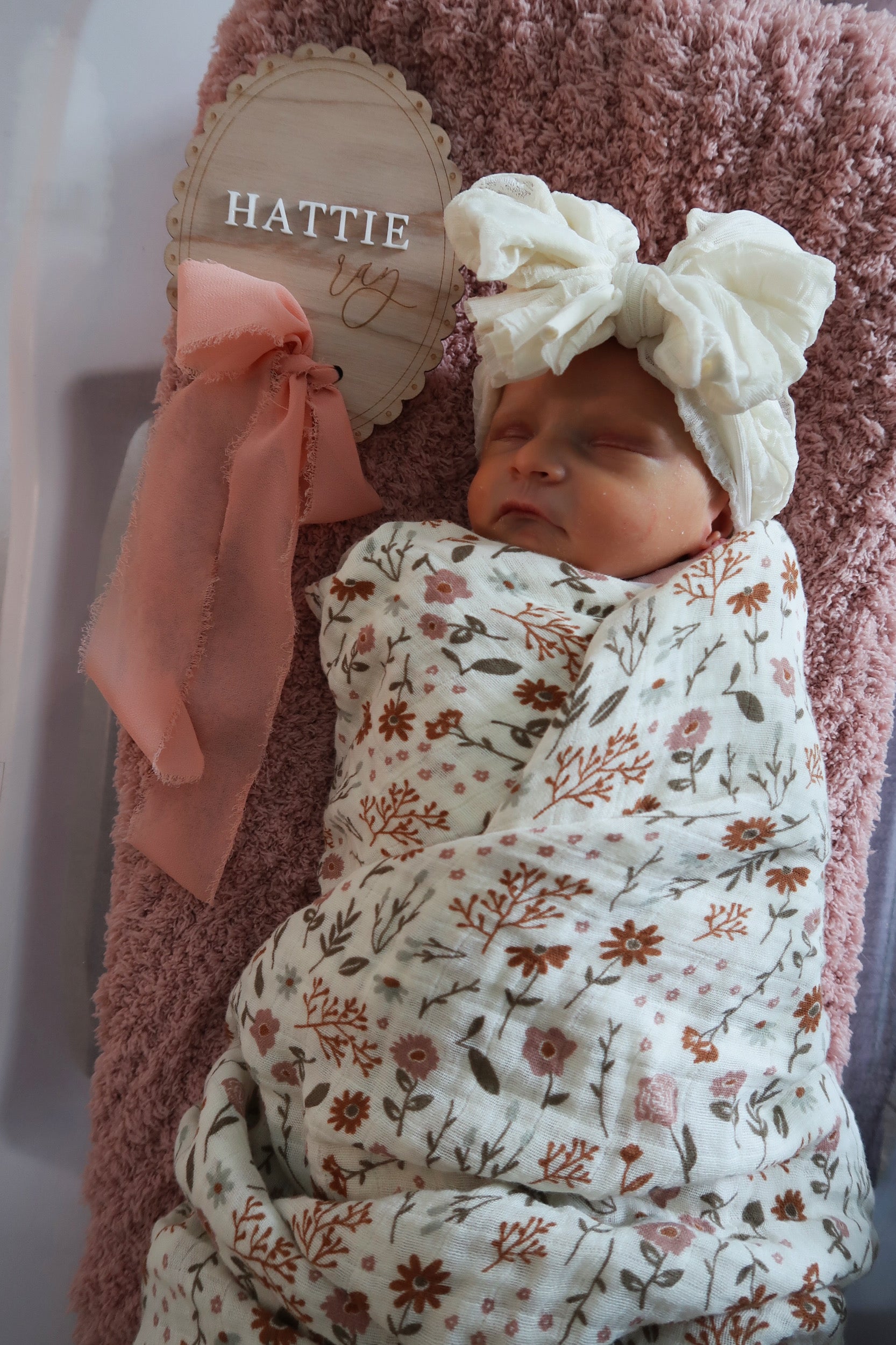 Newborn baby wrapped in a floral swaddle with a white bow headband, sitting on a pink blanket.