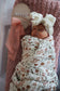 Newborn baby wrapped in a floral swaddle with a white bow headband, sitting on a pink blanket.