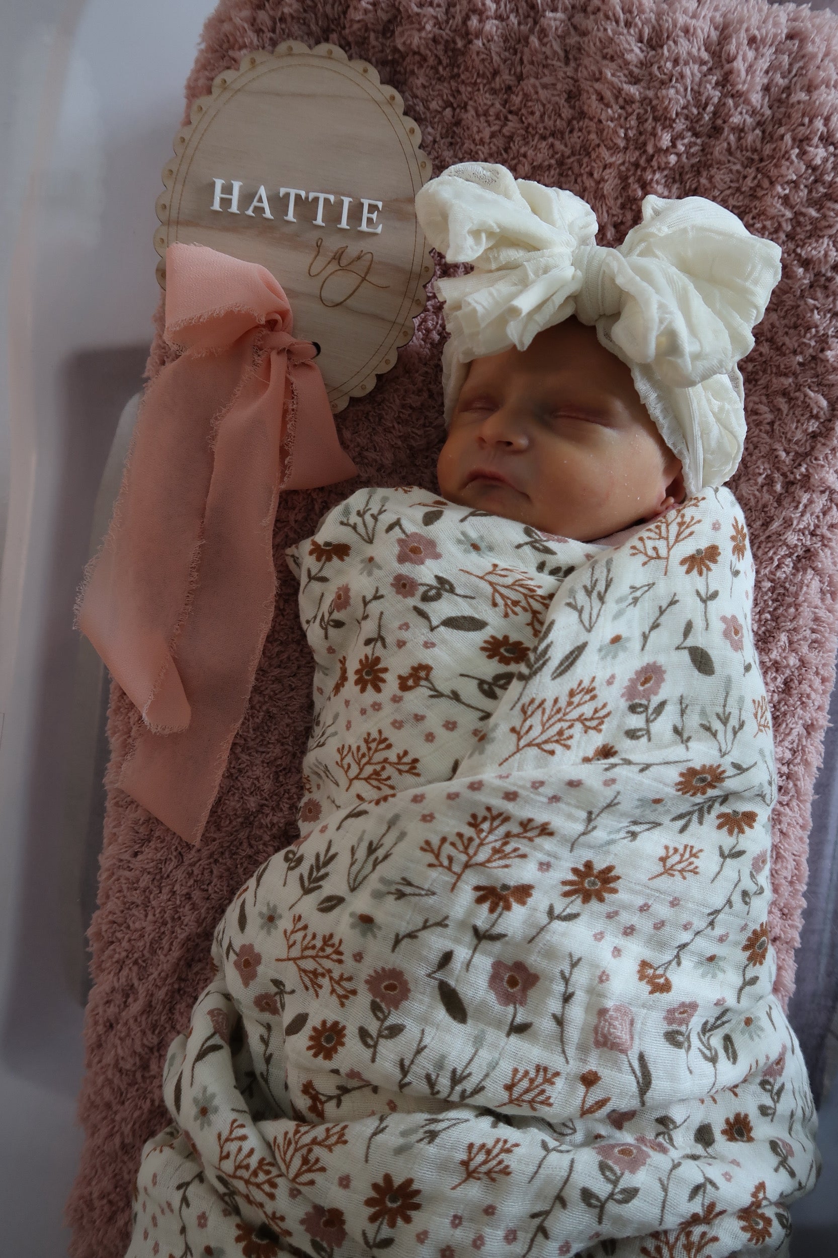 Newborn baby wrapped in a floral swaddle with a headband, sitting on a pink blanket.