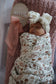 Newborn baby wrapped in a floral swaddle with a headband, sitting on a pink blanket.